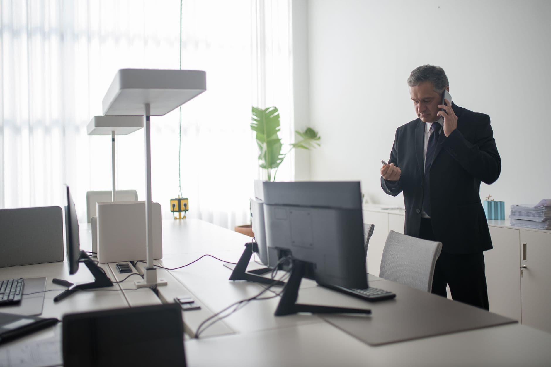 Senior businessman in a black suit takes a call in a sunlit modern office space.