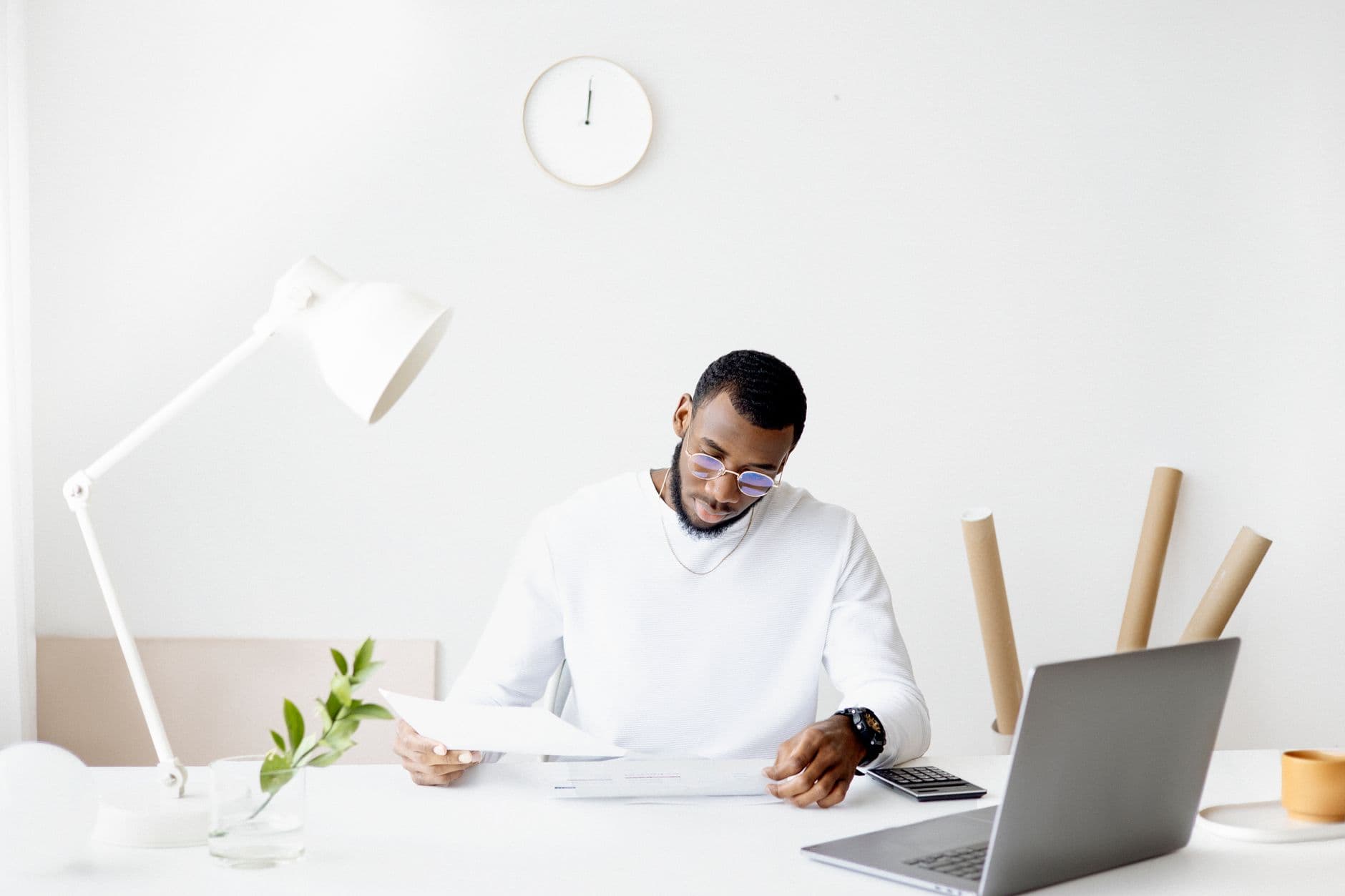 A professional adult working intently at a minimalist desk with a laptop, papers, and a lamp.