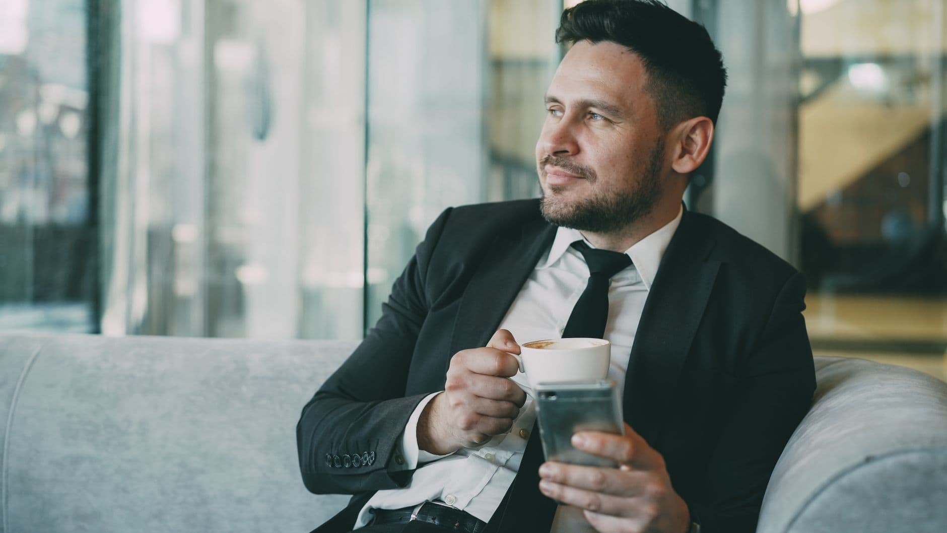A man in a suit relaxing with a coffee while using a smartphone indoors.