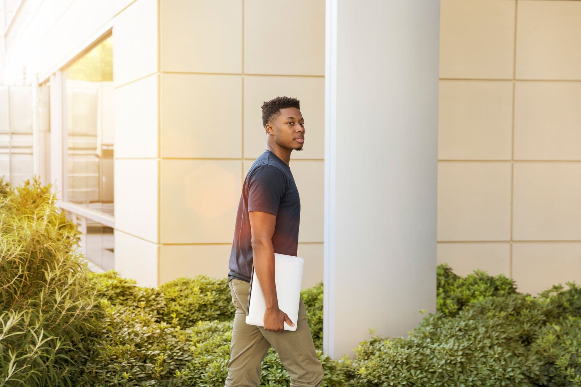 A young man walks outdoors holding a laptop, surrounded by greenery and modern architecture.