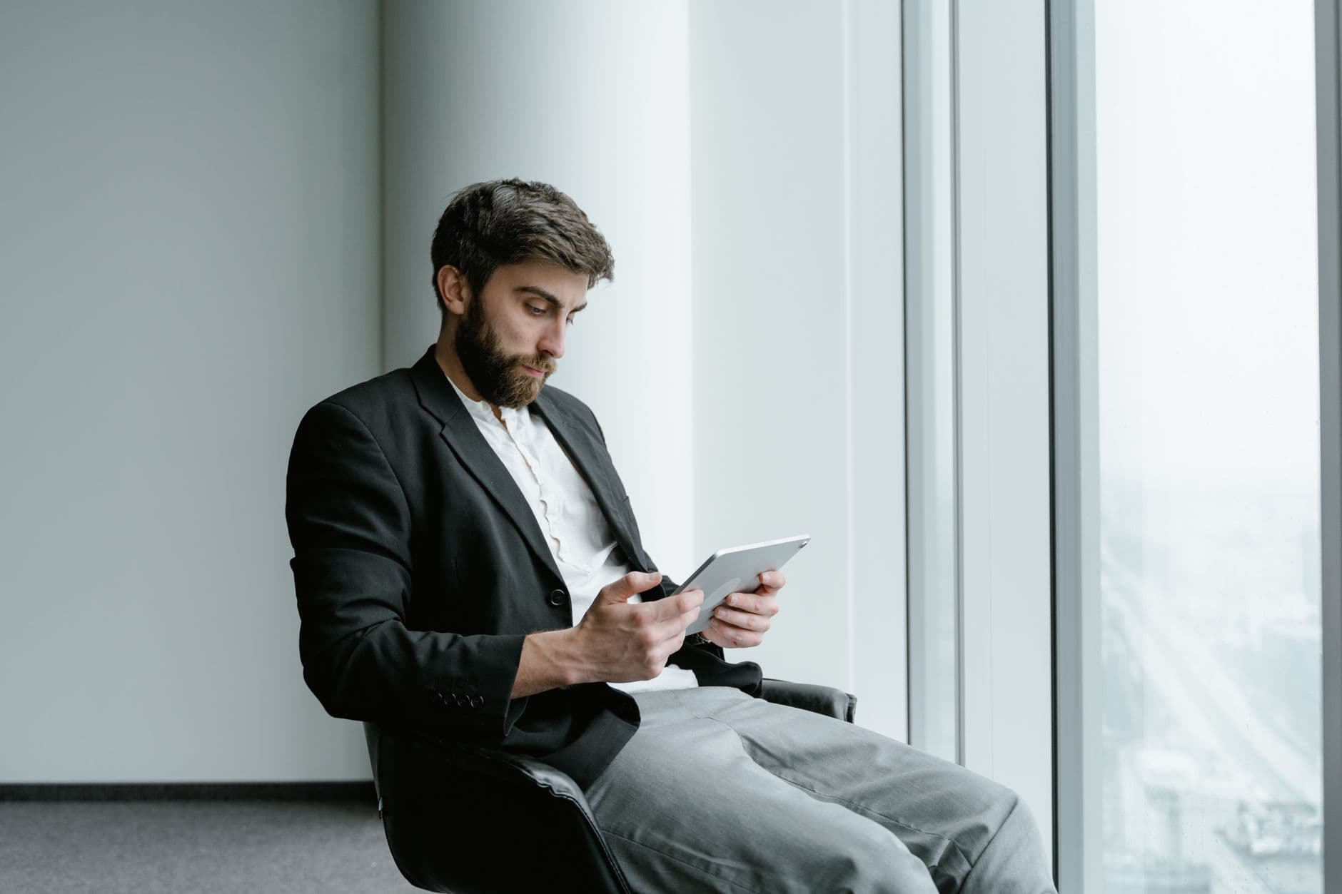 Bearded businessman in black suit using tablet by a large glass window in an office setting.